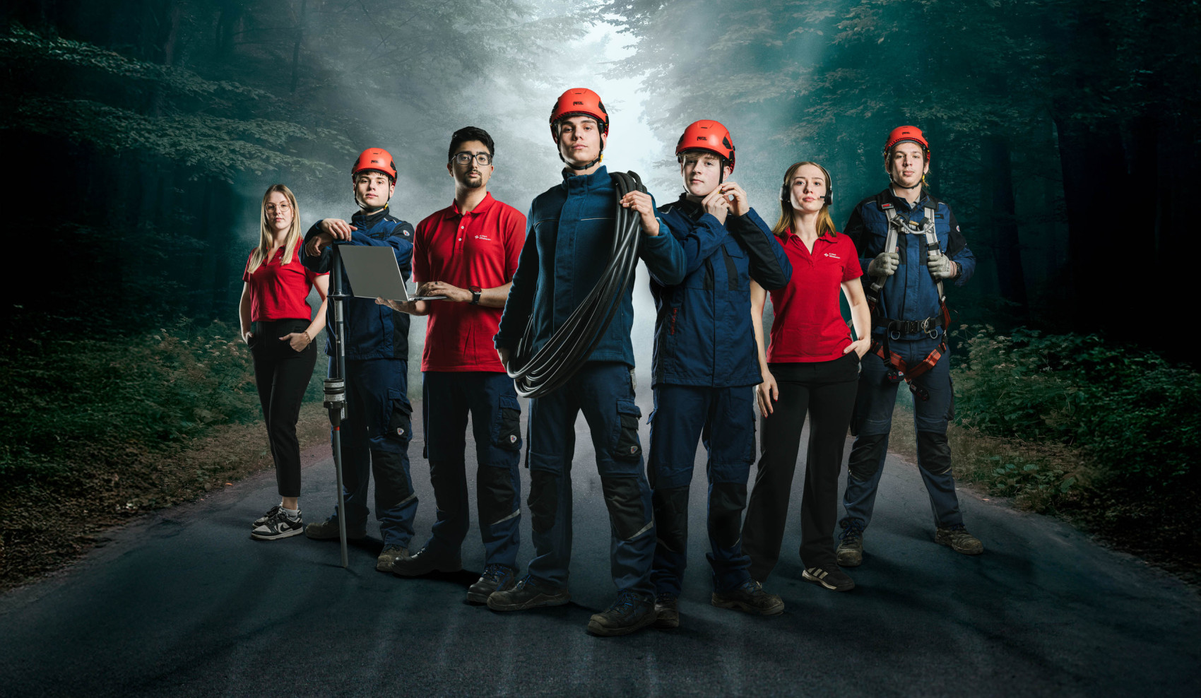 Ein Team von sieben Techniker:innen und Servicemitarbeitenden steht selbstbewusst auf einer Waldstraße, teils in Arbeitskleidung mit Helm, teils in rotem Poloshirt