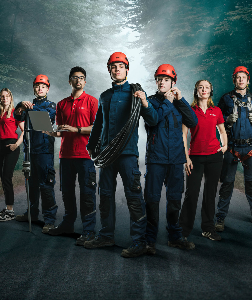 Ein Team von sieben Techniker:innen und Servicemitarbeitenden steht selbstbewusst auf einer Waldstraße, teils in Arbeitskleidung mit Helm, teils in rotem Poloshirt