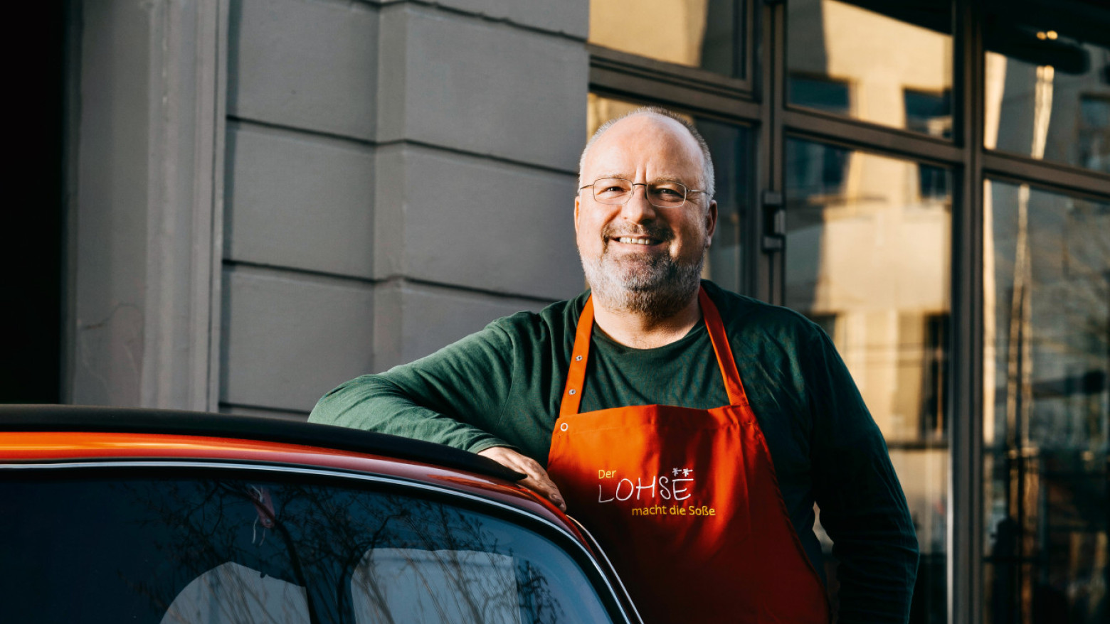 Mann mit oranger Schürze lehnt lächelnd an einem roten Oldtimer vor einem Restaurant 