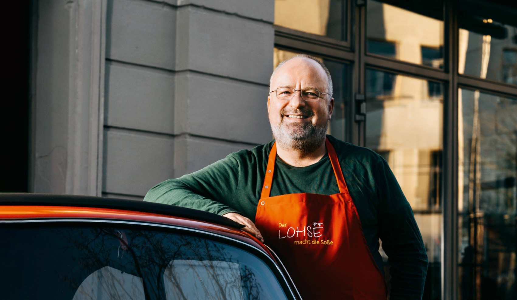 Mann mit oranger Schürze lehnt lächelnd an einem roten Oldtimer vor einem Restaurant 