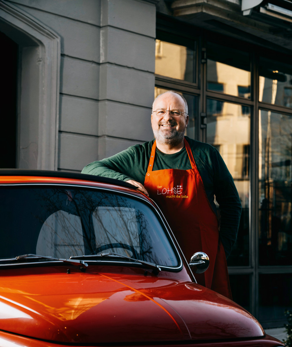 Mann mit oranger Schürze lehnt lächelnd an einem roten Oldtimer vor einem Restaurant 