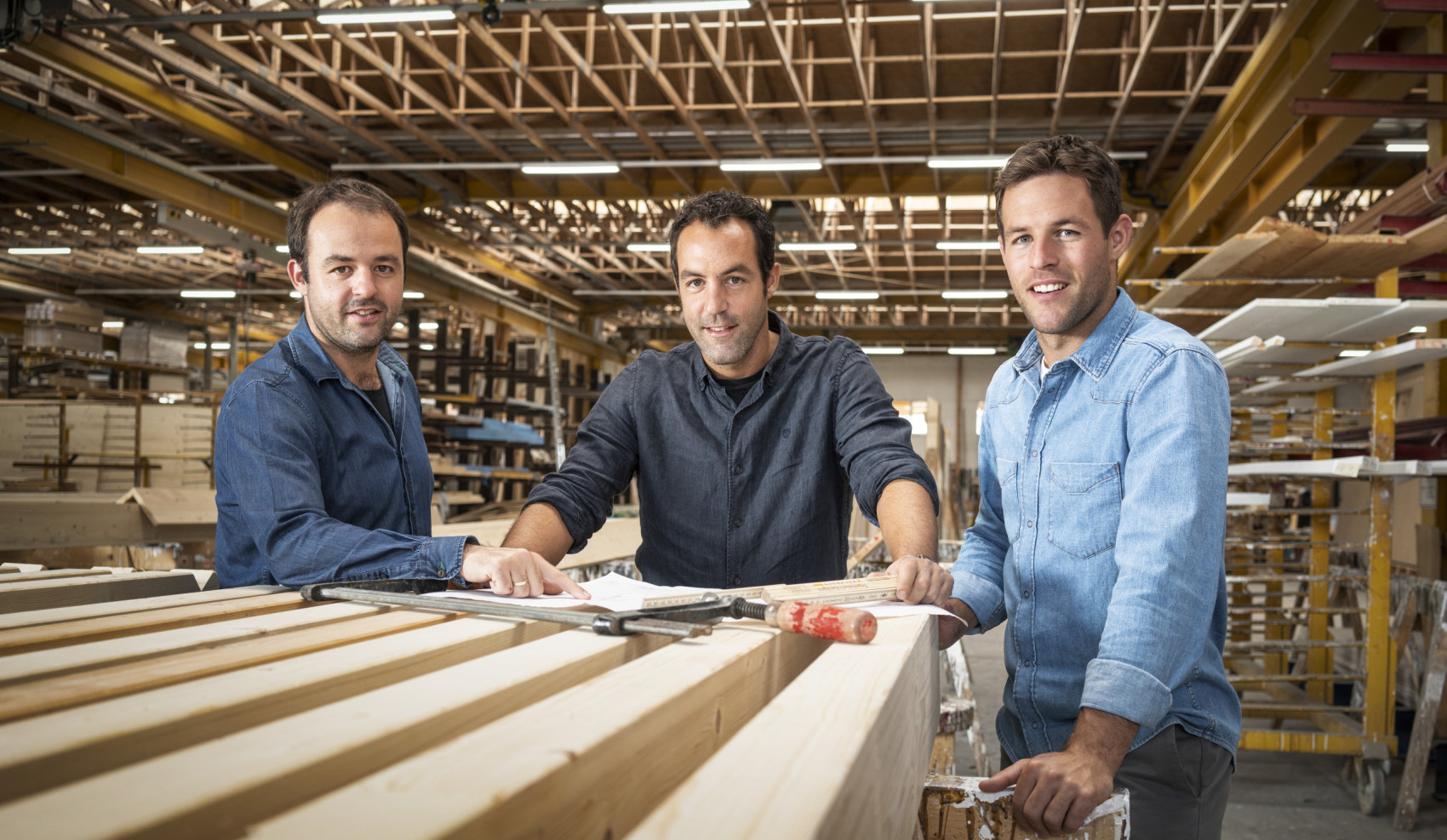 Drei Männer stehen in einer modernen Werkhalle an einem Tisch mit Holzbalken