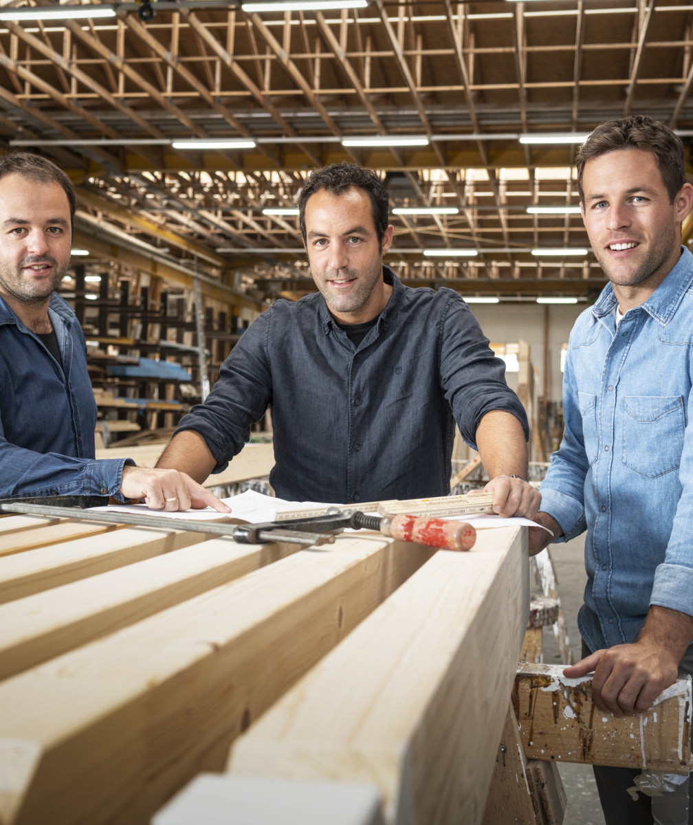 Drei Männer stehen in einer modernen Werkhalle an einem Tisch mit Holzbalken
