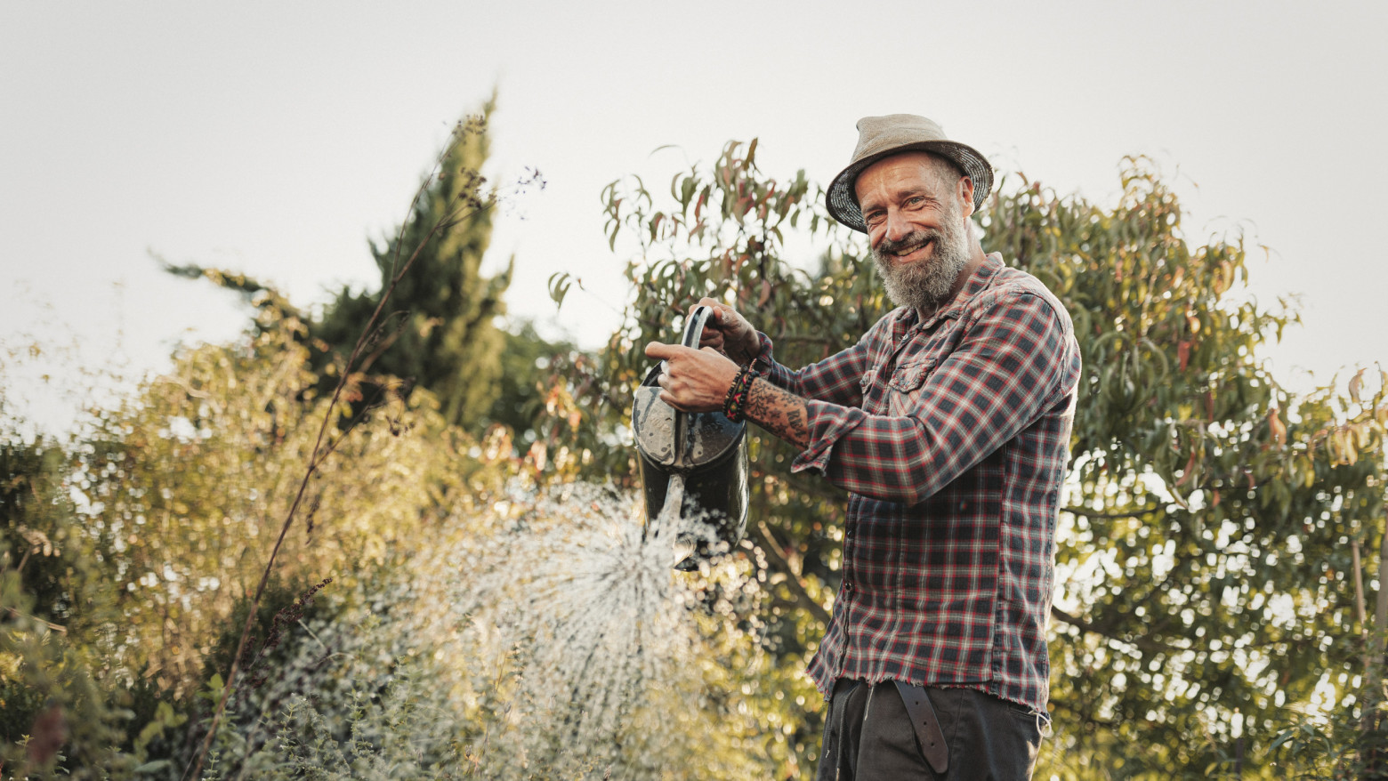 Älterer Mann mit Hut gießt lachend Pflanzen im Garten