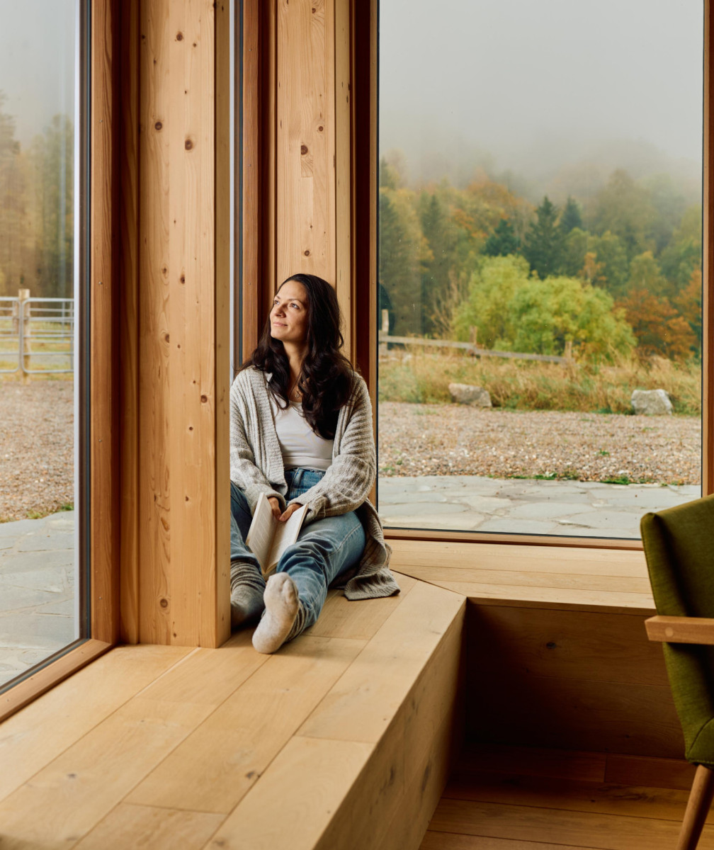 Junge Frau sitzt auf Holzbank am Fenster eines Holzhauses und blickt aus dem Fenster