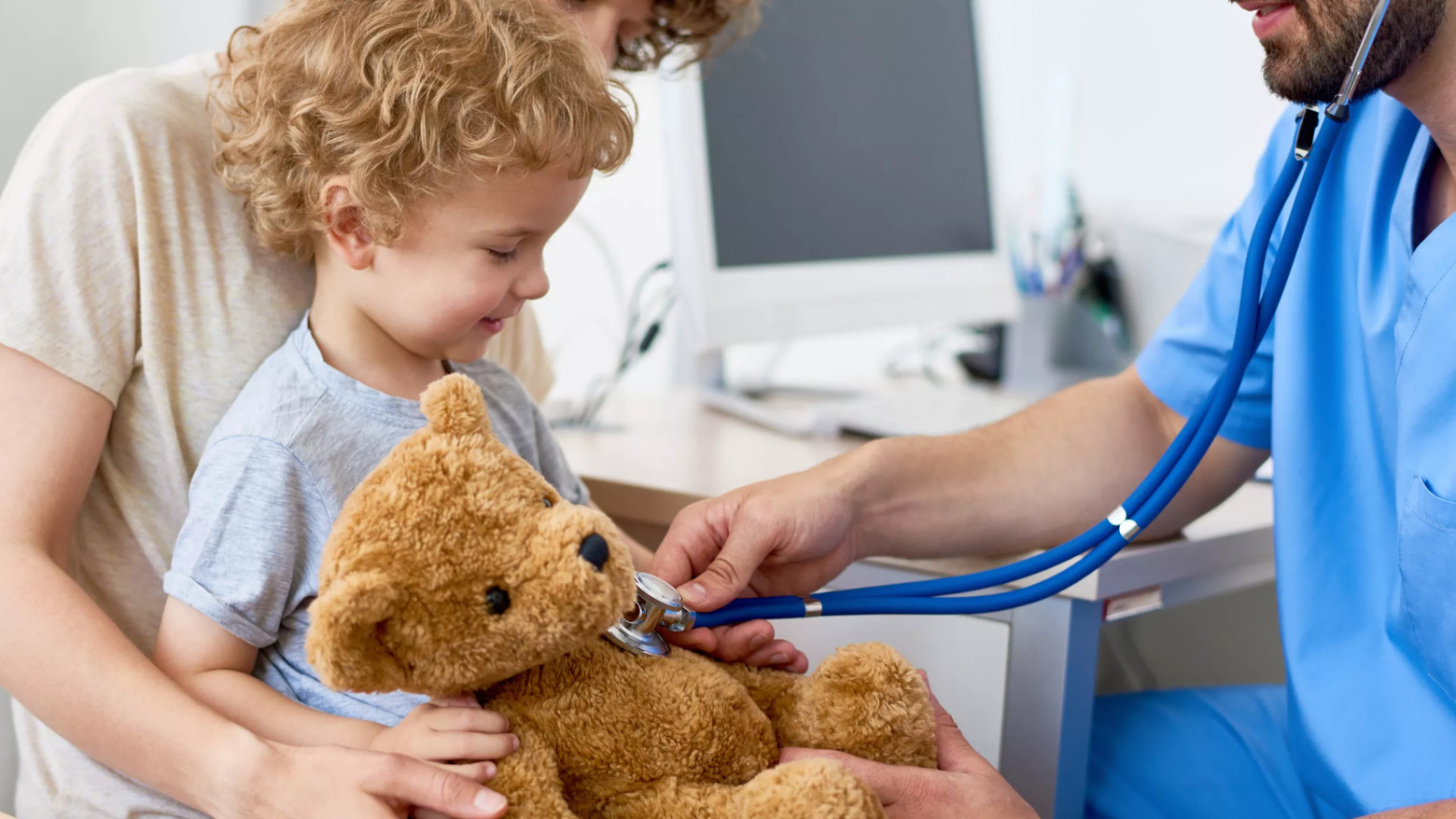 Kind sitzt auf dem Schoß eines Elternteils und lacht, während ein Arzt mit Stethoskop den Plüsch-Teddy abhört