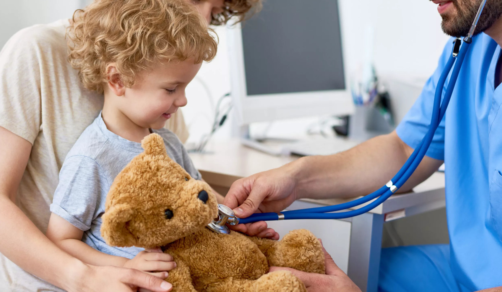 Kind sitzt auf dem Schoß eines Elternteils und lacht, während ein Arzt mit Stethoskop den Plüsch-Teddy abhört