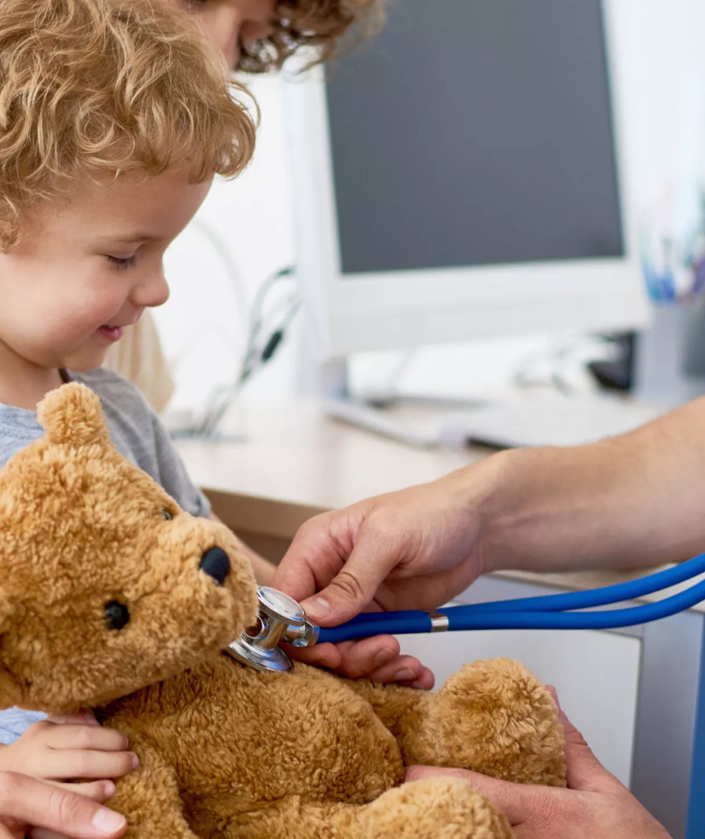 Kind sitzt auf dem Schoß eines Elternteils und lacht, während ein Arzt mit Stethoskop den Plüsch-Teddy abhört