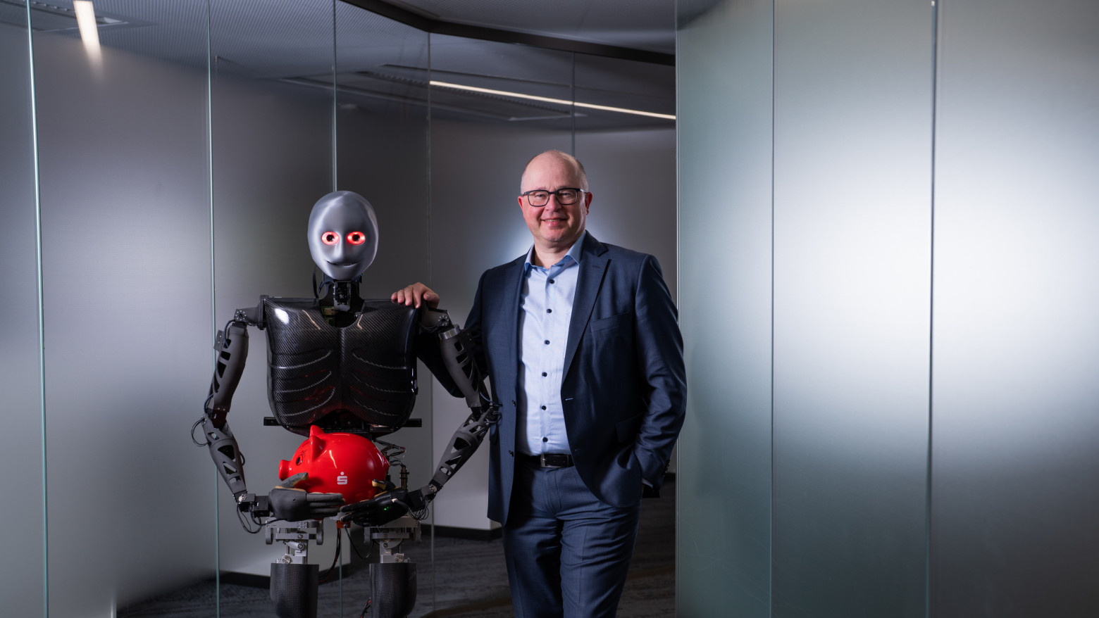 Ein humanoider Roboter hält ein rotes Sparschwein, neben Ihm steht lächelnd ein Mann im Anzug