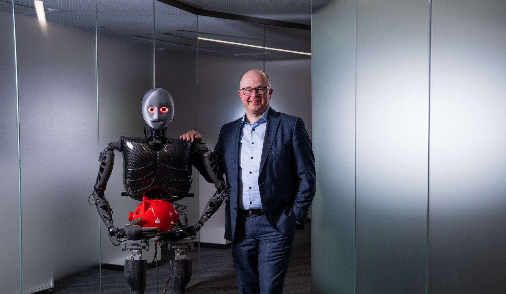 Ein humanoider Roboter hält ein rotes Sparschwein, neben Ihm steht lächelnd ein Mann im Anzug