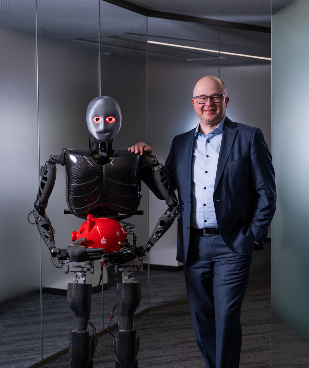 Ein humanoider Roboter hält ein rotes Sparschwein, neben Ihm steht lächelnd ein Mann im Anzug
