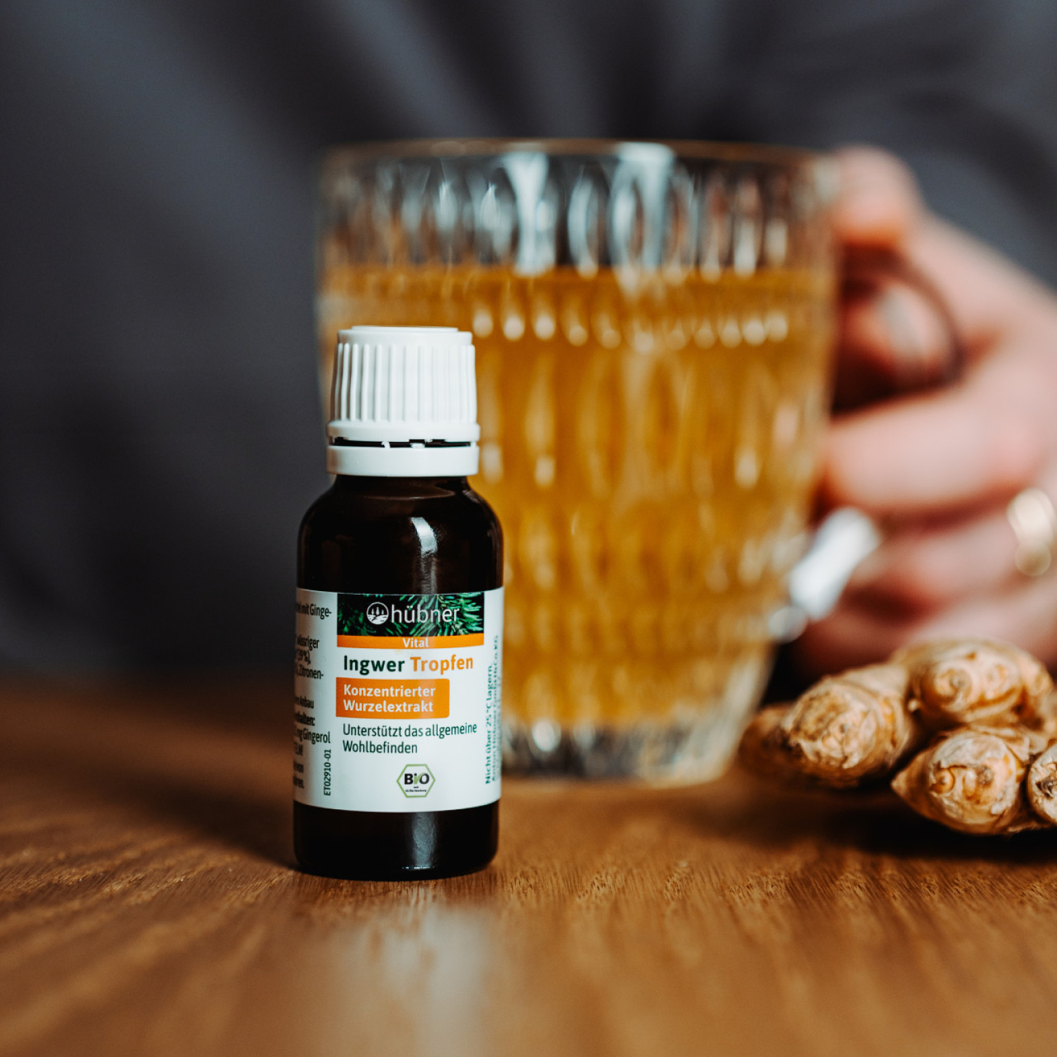 Nahaufnahme einer kleinen Flasche Ingwer-Tropfen auf Holztisch, im Hintergrund eine Hand mit Teeglas und einige frische Ingwerknollen