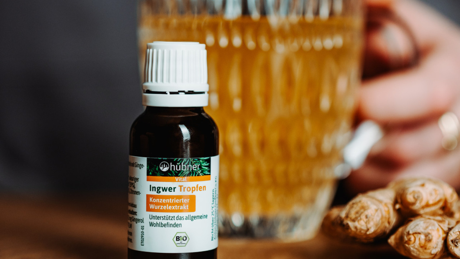 Nahaufnahme einer kleinen Flasche Ingwer-Tropfen auf Holztisch, im Hintergrund eine Hand mit Teeglas und einige frische Ingwerknollen