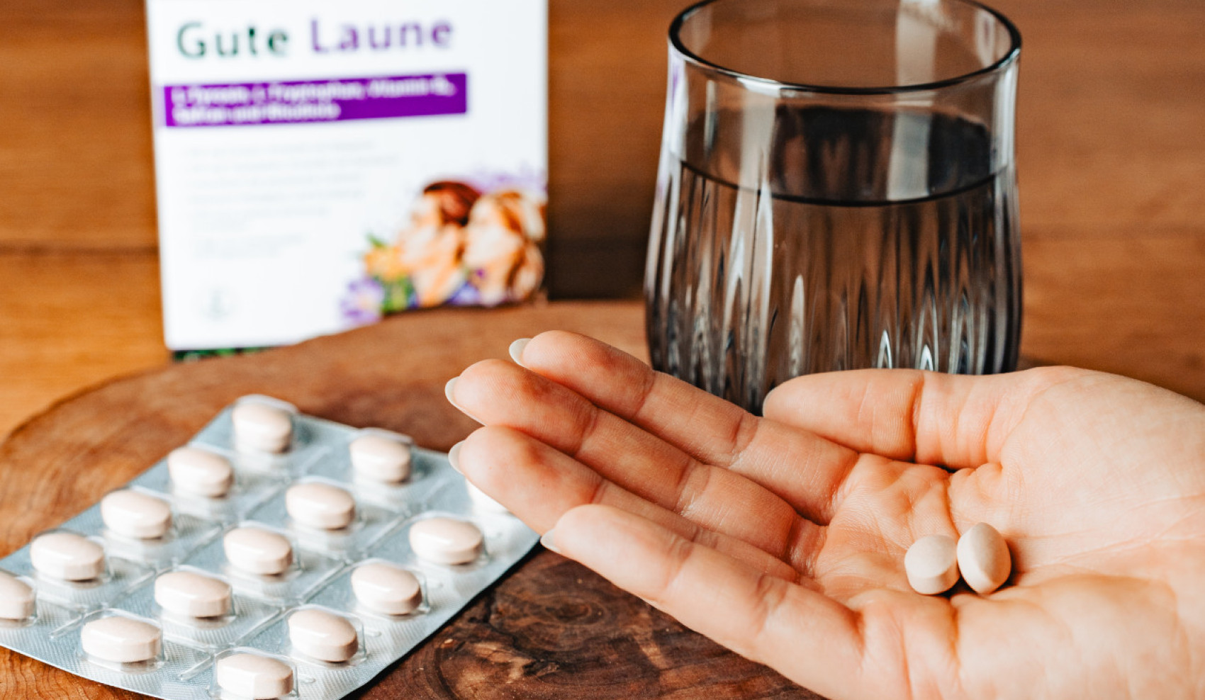 Hand mit Nahrungsergänzungskapseln über Holzbrett, daneben Blisterpackung, Glas Wasser und Verpackung „Gute Laune“ von Hübner