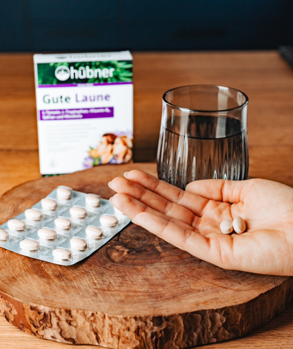 Hand mit Nahrungsergänzungskapseln über Holzbrett, daneben Blisterpackung, Glas Wasser und Verpackung „Gute Laune“ von Hübner