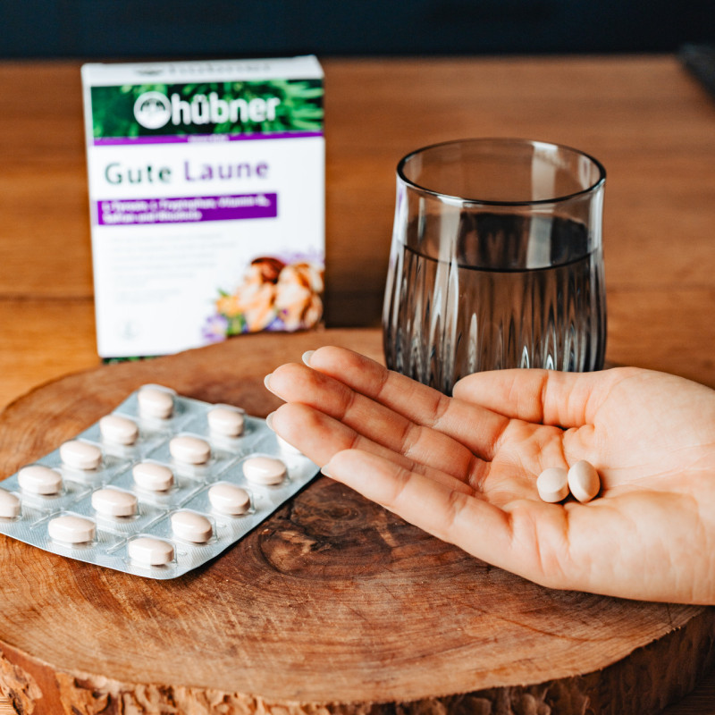 Hand mit Nahrungsergänzungskapseln über Holzbrett, daneben Blisterpackung, Glas Wasser und Verpackung „Gute Laune“ von Hübner