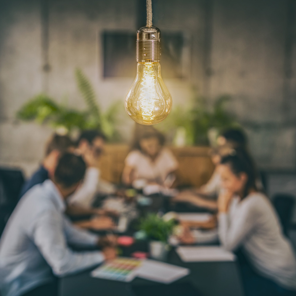 Leuchtende Glühbirne im Fokus, im Hintergrund unscharf eine Gruppe bei einer Besprechung im modernen Büro.