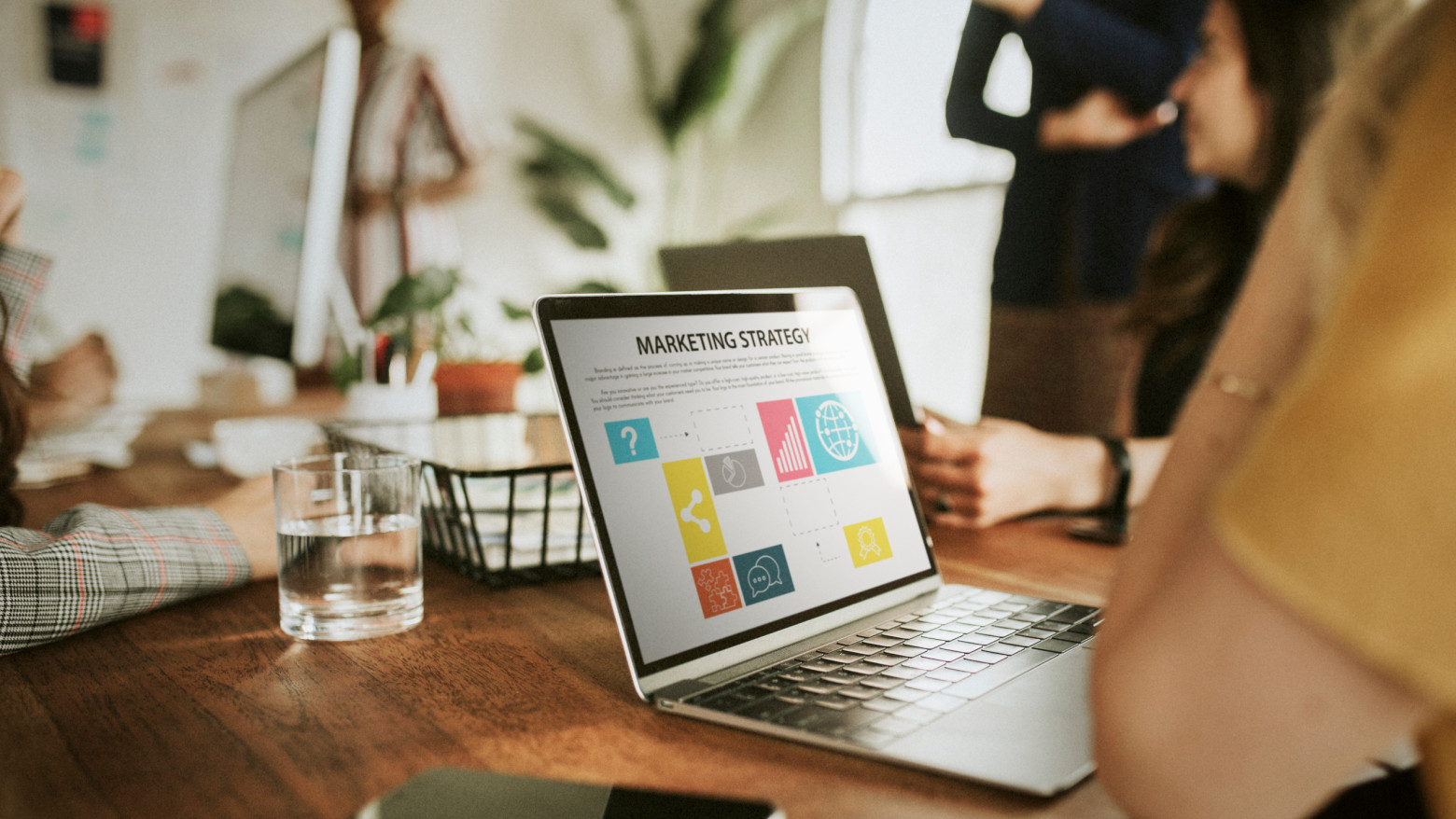 Person sitzt vor Laptop, auf dem Bildschirm eine Präsentation über Marketing Strategy, unscharf mehrere Personen mit Laptops im Hintergrund.