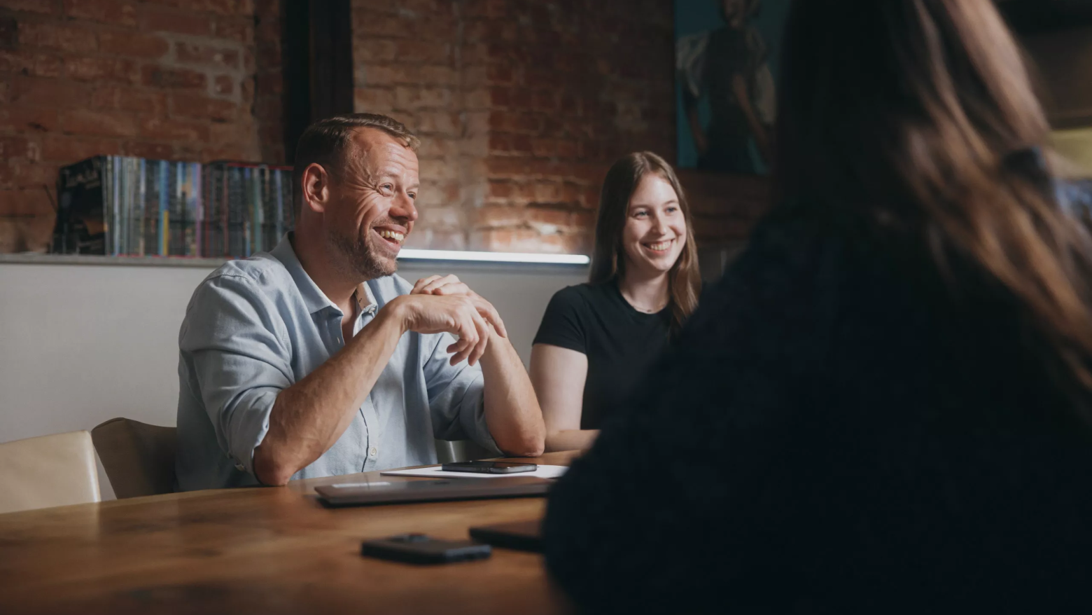 Das Bild zeigt drei Personen in einem Gespräch an einem Konferenztisch in gemütlicher Atmosphäre. Zwei davon – ein Mann in einem hellblauen Hemd und eine junge Frau in einem schwarzen Shirt – lachen herzlich, während sie der dritten Person gegenübersitzen. Die Szene wirkt freundlich, offen und ungezwungen. Im Hintergrund sind eine freigelegte Backsteinwand, Bücher und ein Kunstposter zu sehen, was dem Raum ein kreatives Flair verleiht.